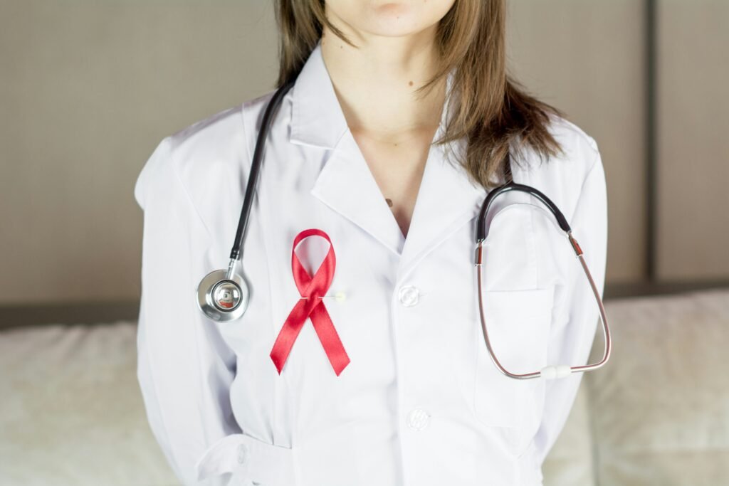 Close-up of a female doctor wearing a white lab coat with a red HIV/AIDS awareness ribbon pinned to it and a stethoscope around her neck, symbolizing support for HIV research and healthcare.