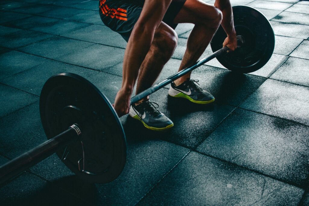 Close‑up of a person’s legs and hands as they grip a loaded barbell on a gym floor, preparing to lift.