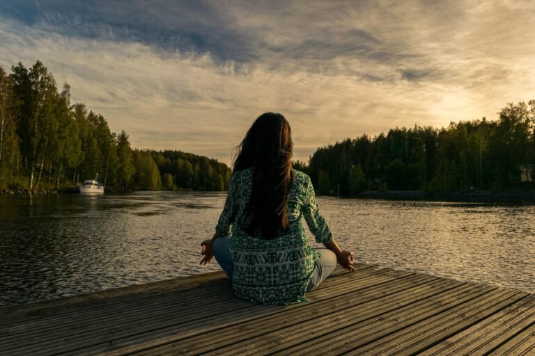 A person seated cross‑legged in a meditative yoga pose on a wooden dock overlooking a calm lake at sunset, with trees lining the shore and a small boat moored nearby.