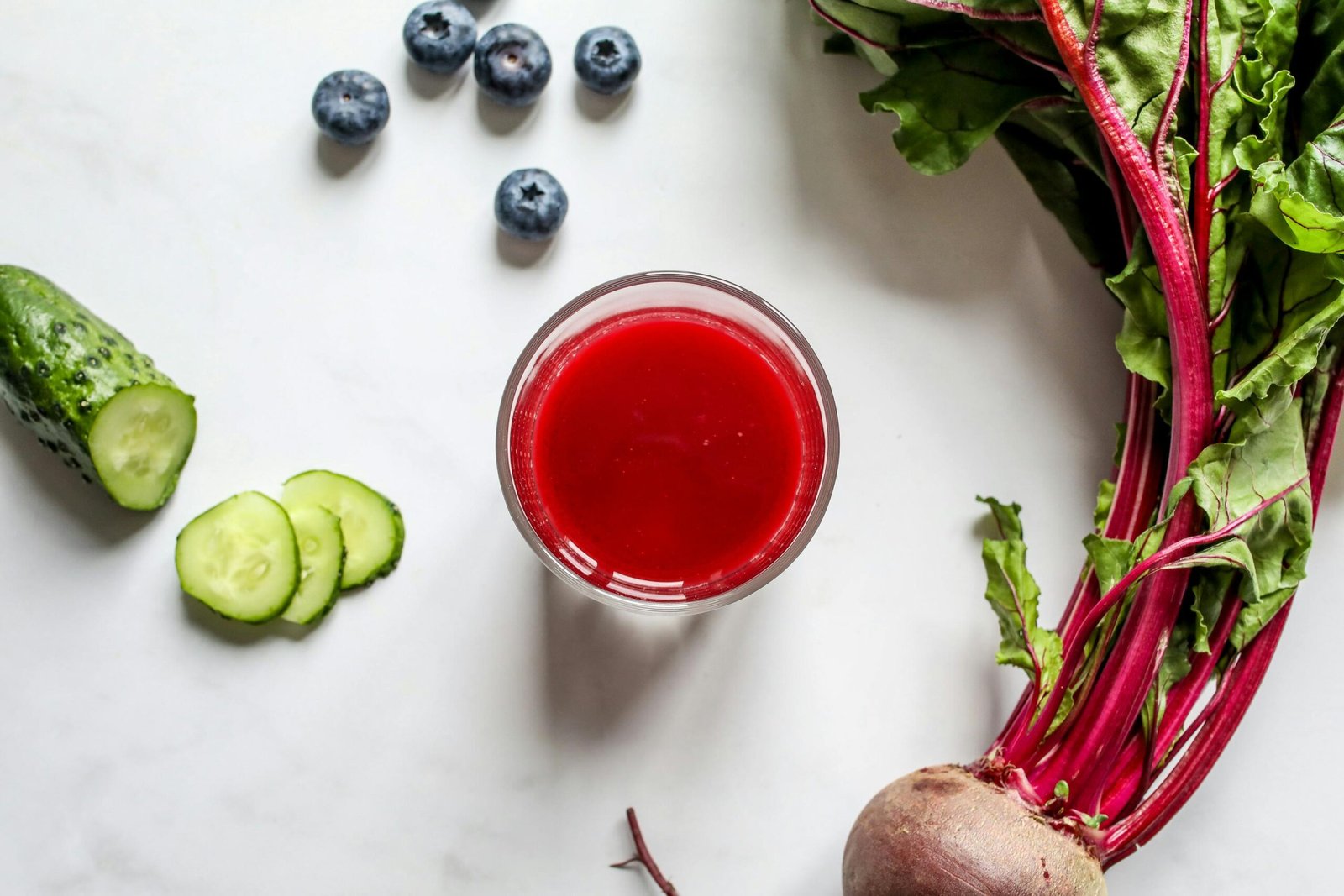 Red Juice in Clear Drinking Glass, Beetroot, Sliced Cucumber and Blueberries on White Table.