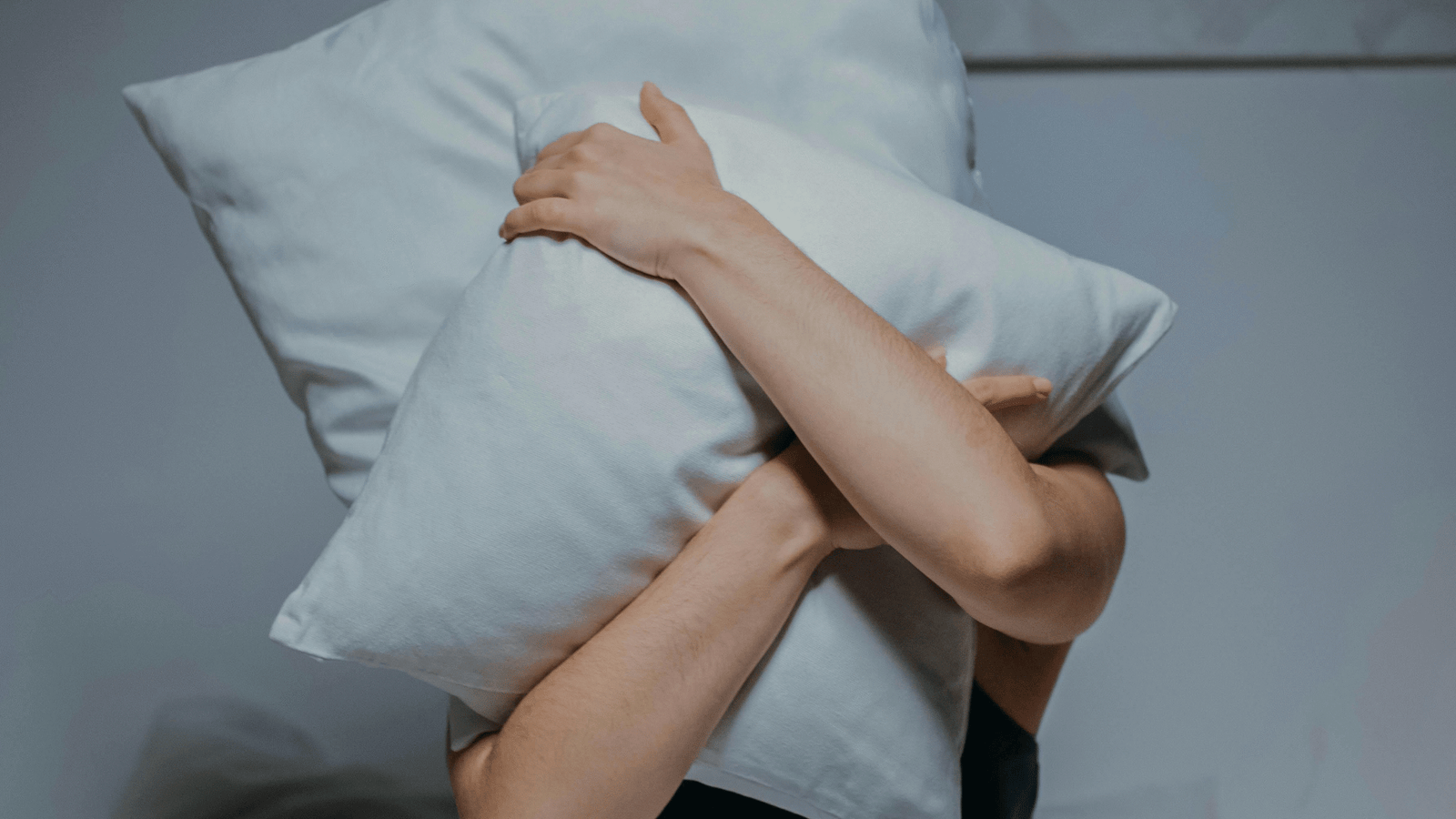 A person sits in bed, covering their face with a pillow in a minimalist bedroom.
