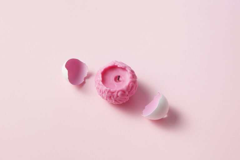 A unique pink candle shaped like a brain surrounded by white eggshells on a light pink background.