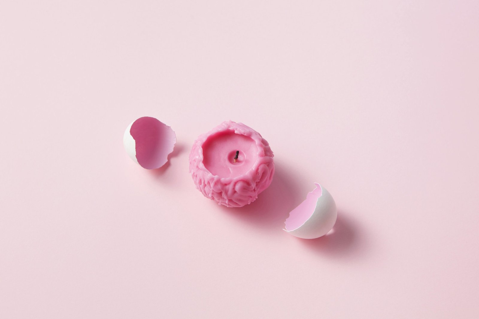 A unique pink candle shaped like a brain surrounded by white eggshells on a light pink background.