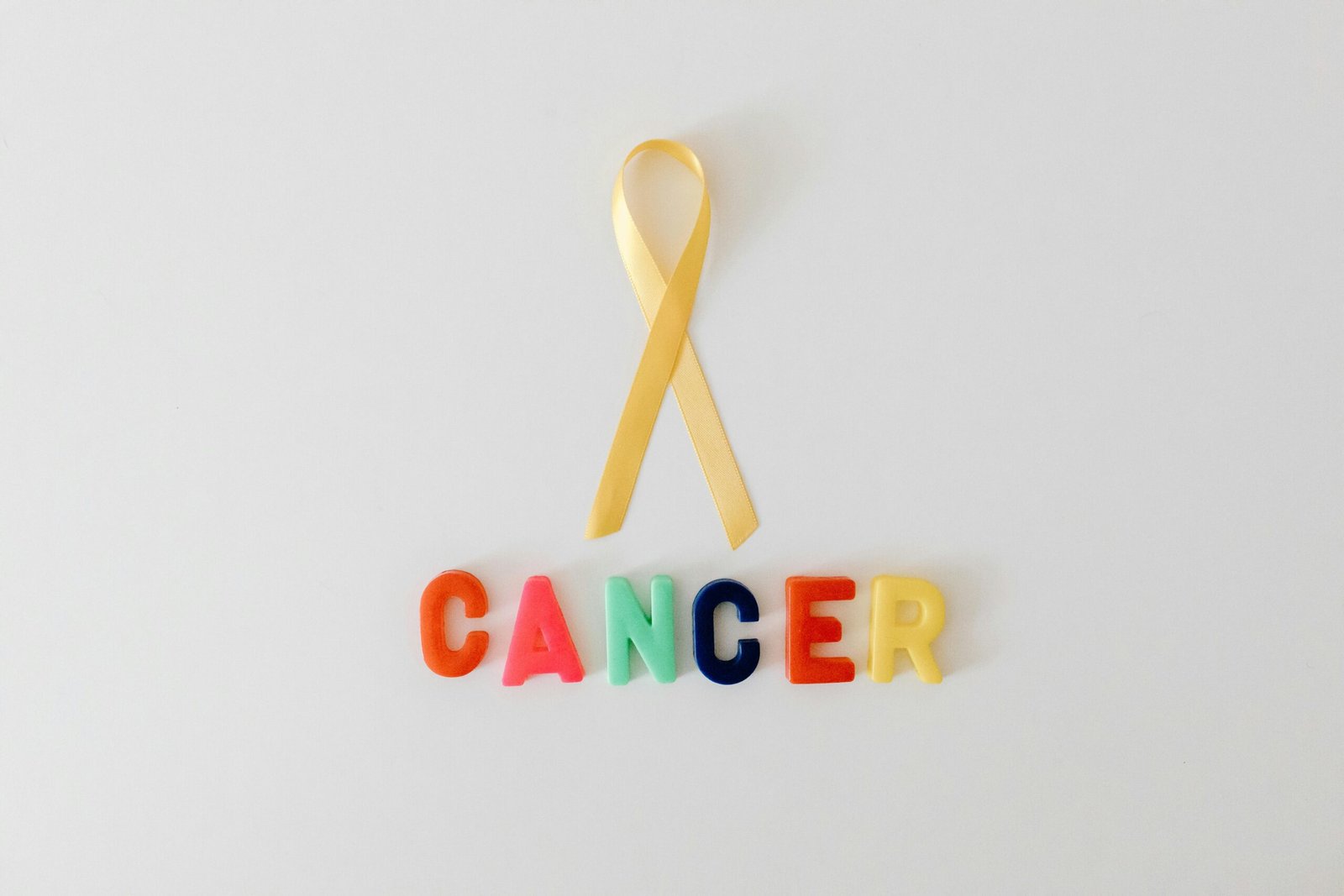 Yellow cancer awareness ribbon with colorful letters spelling 'cancer' on white background.
