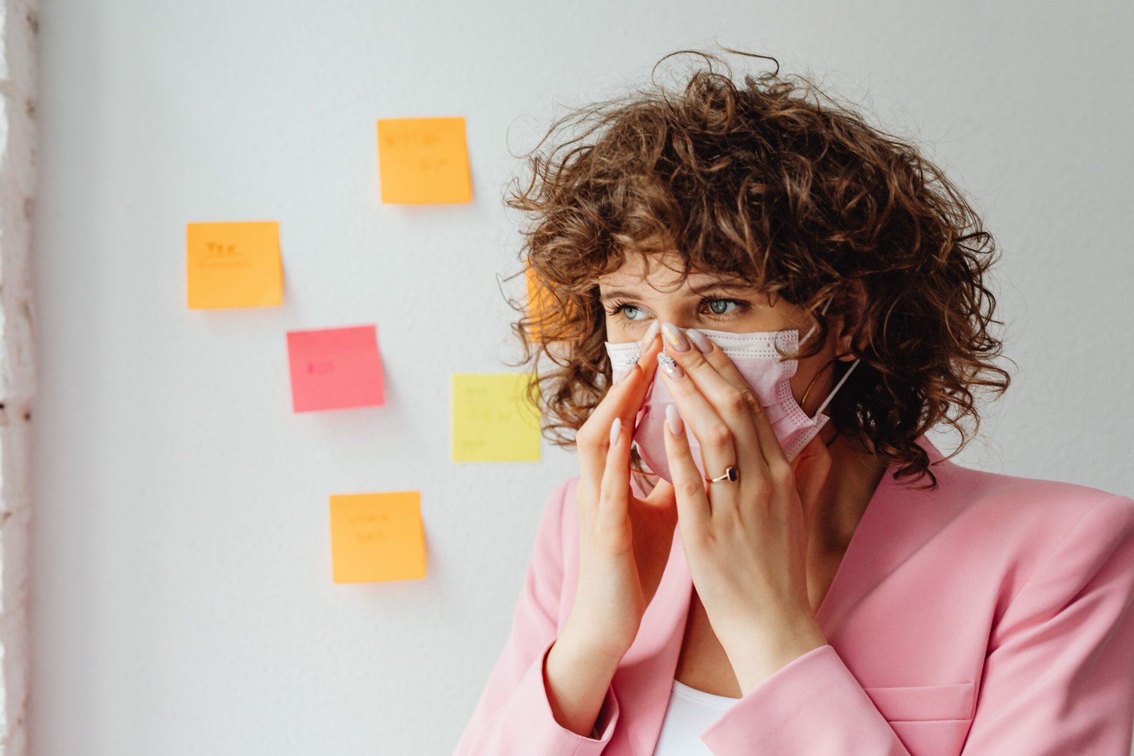Thoughtful woman with curly hair in a pink blazer wearing a face mask indoors.